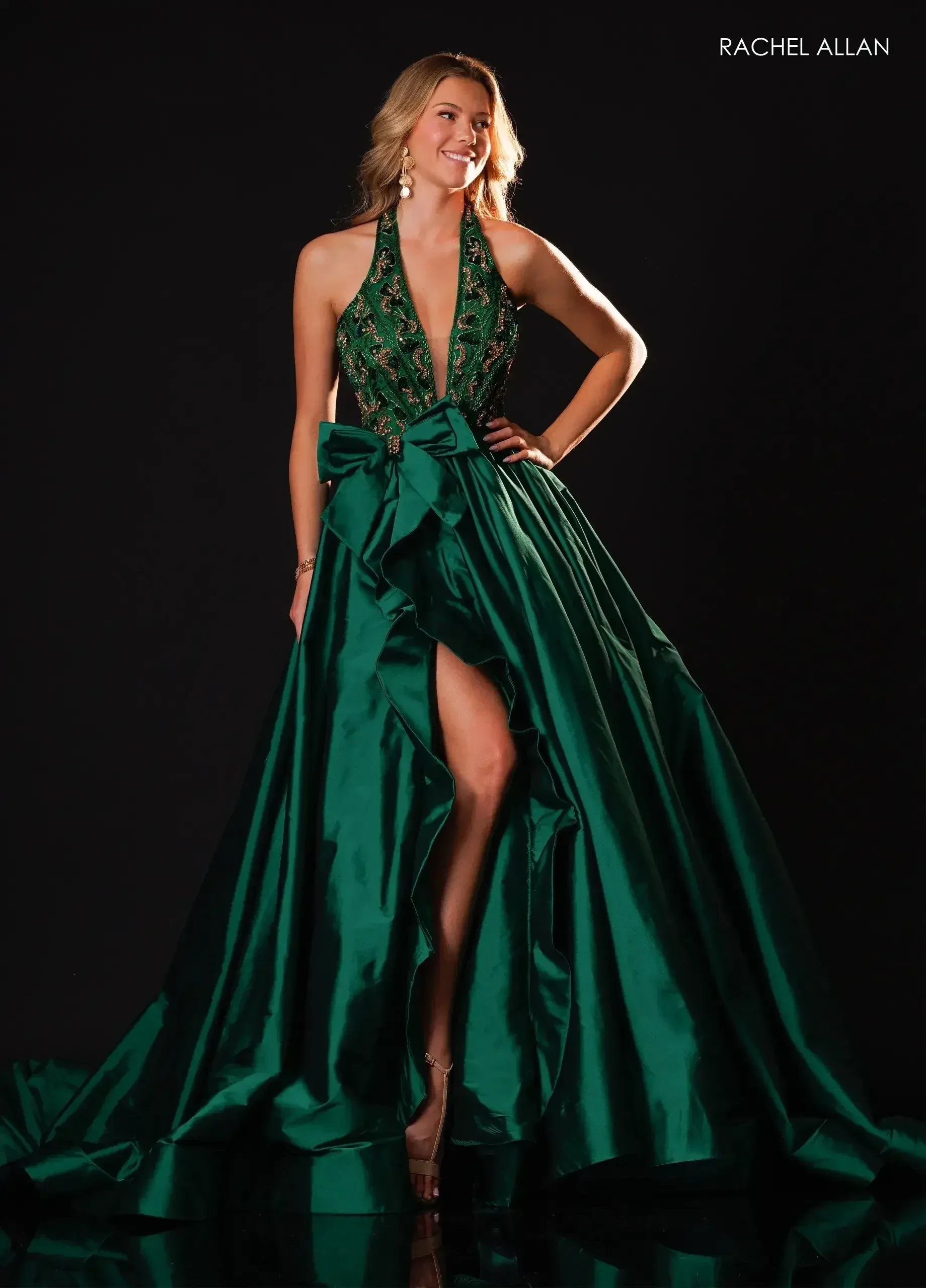 Model wearing an emerald green Rachel Allan halter gown with an embellished bodice, oversized bow, high slit, and full satin skirt.